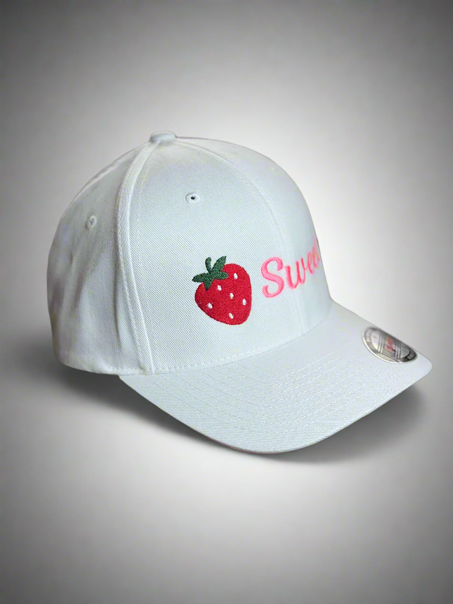 A white baseball cap with the word “Sweet” in pink next to an embroidered red strawberry, viewed from a side angle.