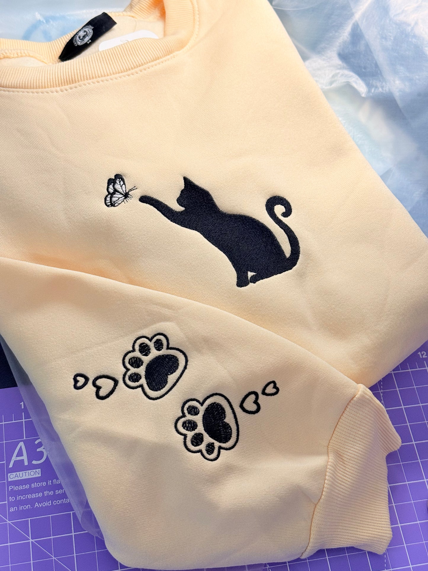 Closeup sleeve view of Unisex light beige hoodie with embroidered chest design featuring a Cat Caught in a Moment with Butterfly