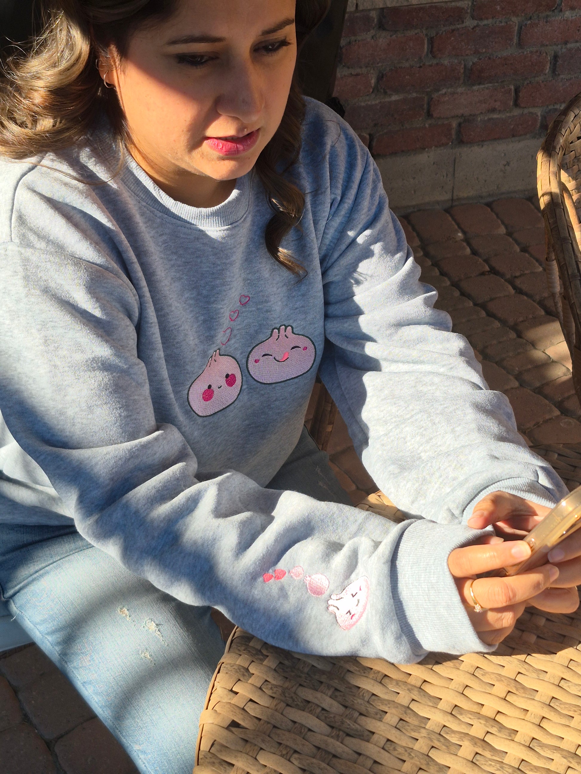 Person wearing a grey Kawaii Dumpling embroidered unisex sweatshirt, showing sleeve detail with pink heart dumpling embroidery.