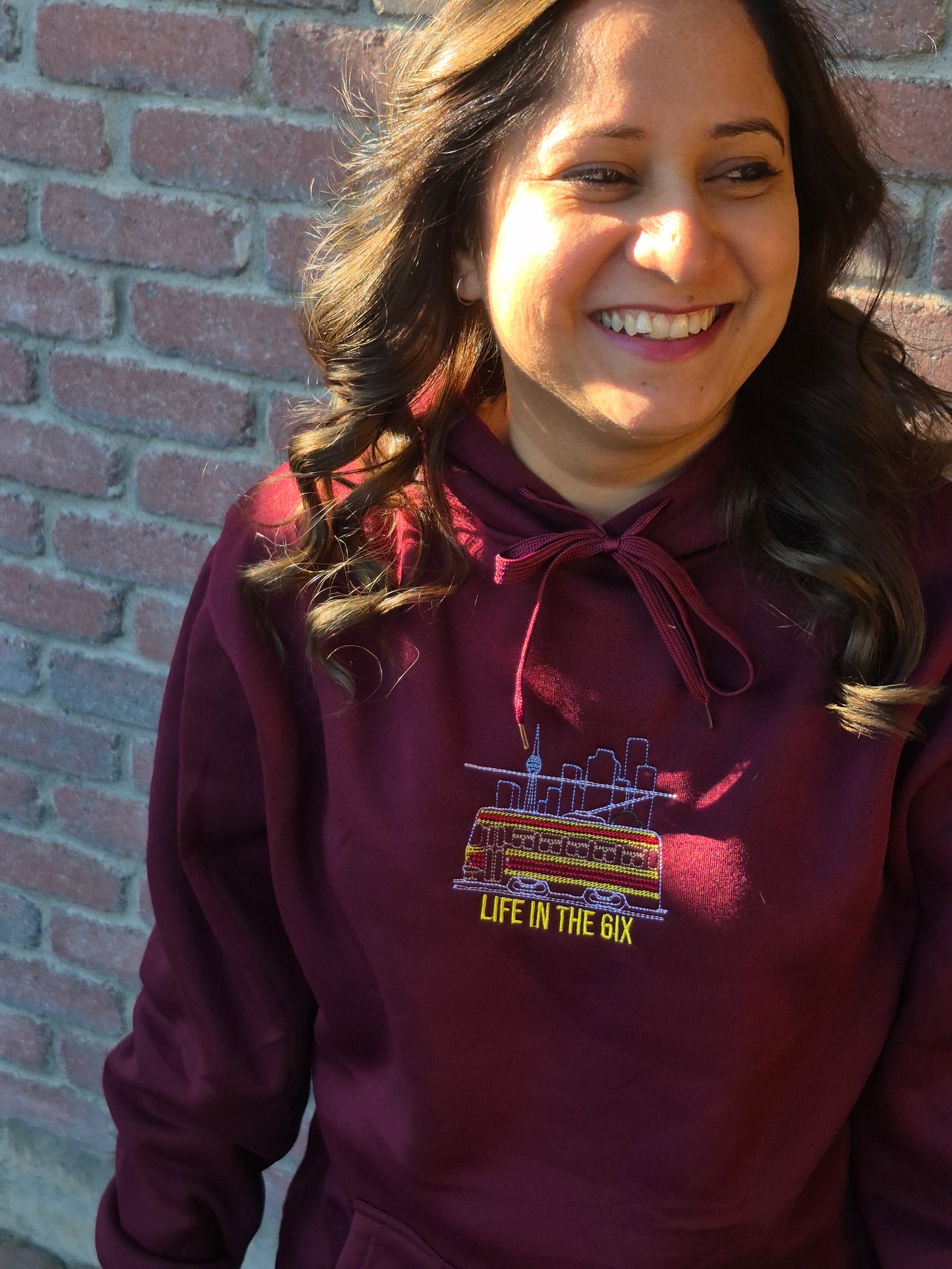 Angled close-up of a person wearing a wine-red Toronto Skyline Streetcar embroidered hoodie highlighting the colorful streetcar and CN Tower graphic with “Life in the 6ix.”