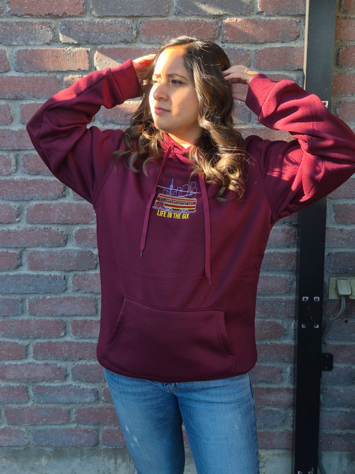 Person wearing a wine-red Toronto Skyline Streetcar embroidered hoodie, featuring CN Tower and “Life in the 6ix” chest design.