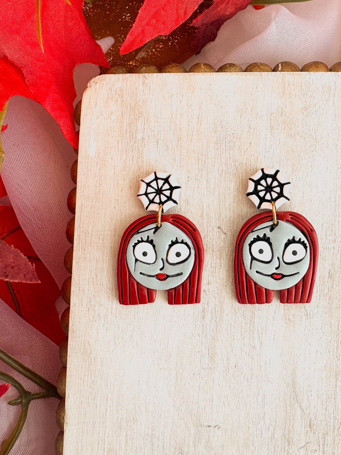 Pair of Halloween Dangle Earrings, featuring polymer clay charms of a cartoon girl with red hair, gray face, and expressive eyes, hanging from white and black spiderweb studs