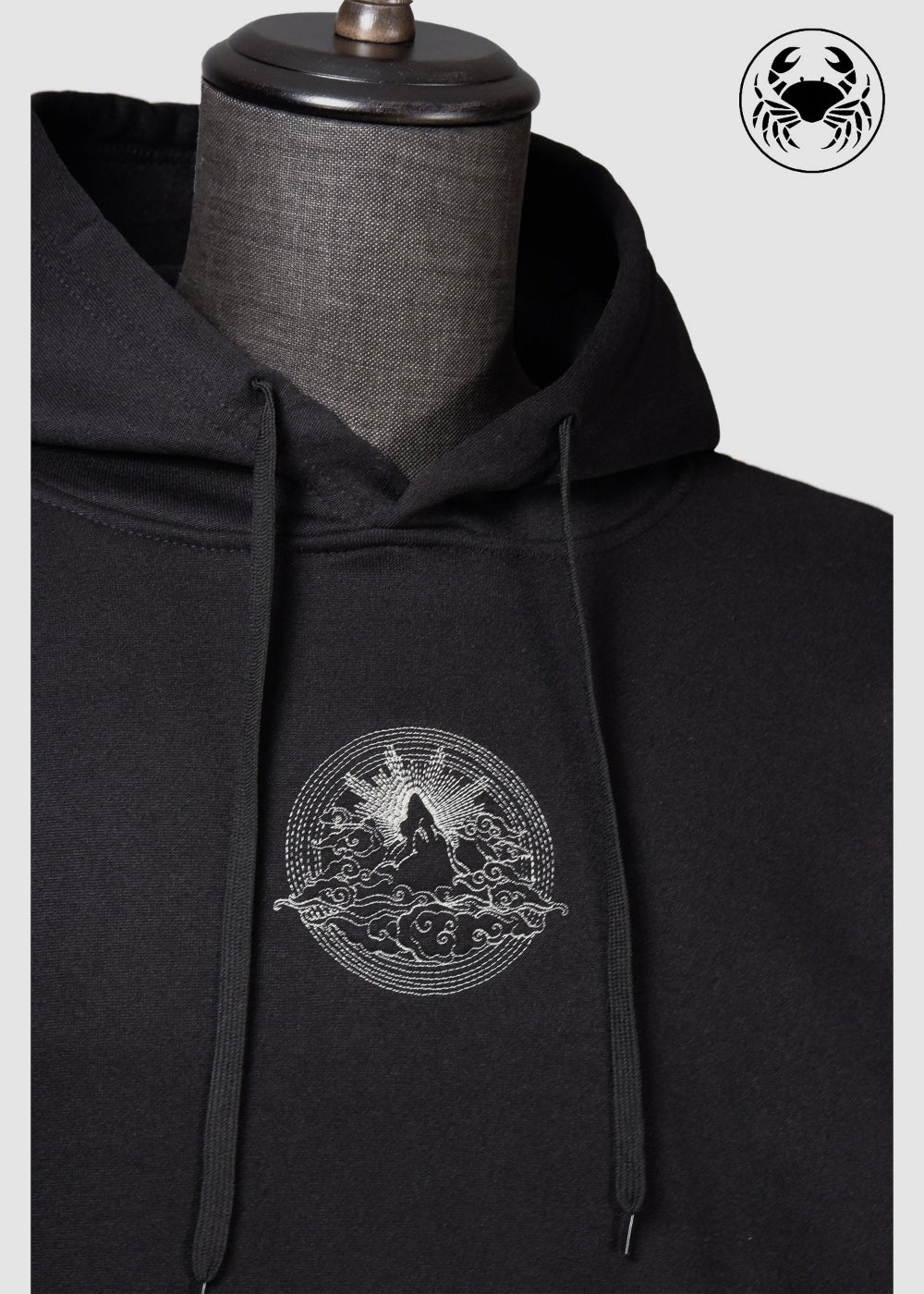 Angled view of Unisex black fleece hoodie featuring a front kangaroo pocket, adjustable drawstring hood, and an embroidered  mountain and sun rising design 