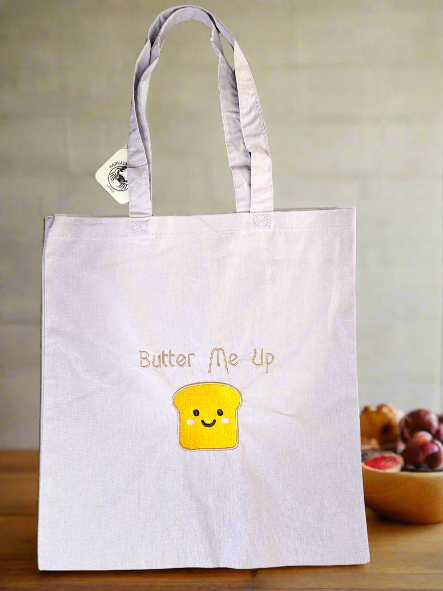 Light purple cotton tote bag with embroidered “Butter Me Up” and smiling toast illustration