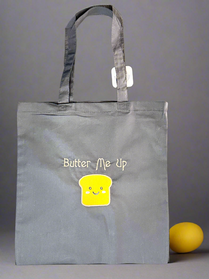 Charcoal grey cotton tote bag with embroidered “Butter Me Up” and yellow smiling toast