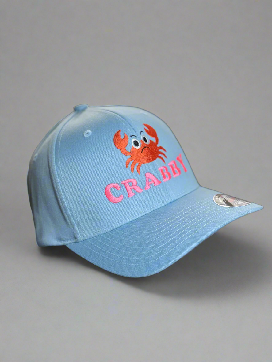 Light blue baseball cap with pink "CRABBY" text and a red cartoon crab embroidery on the front. Playful and coastal-themed streetwear design