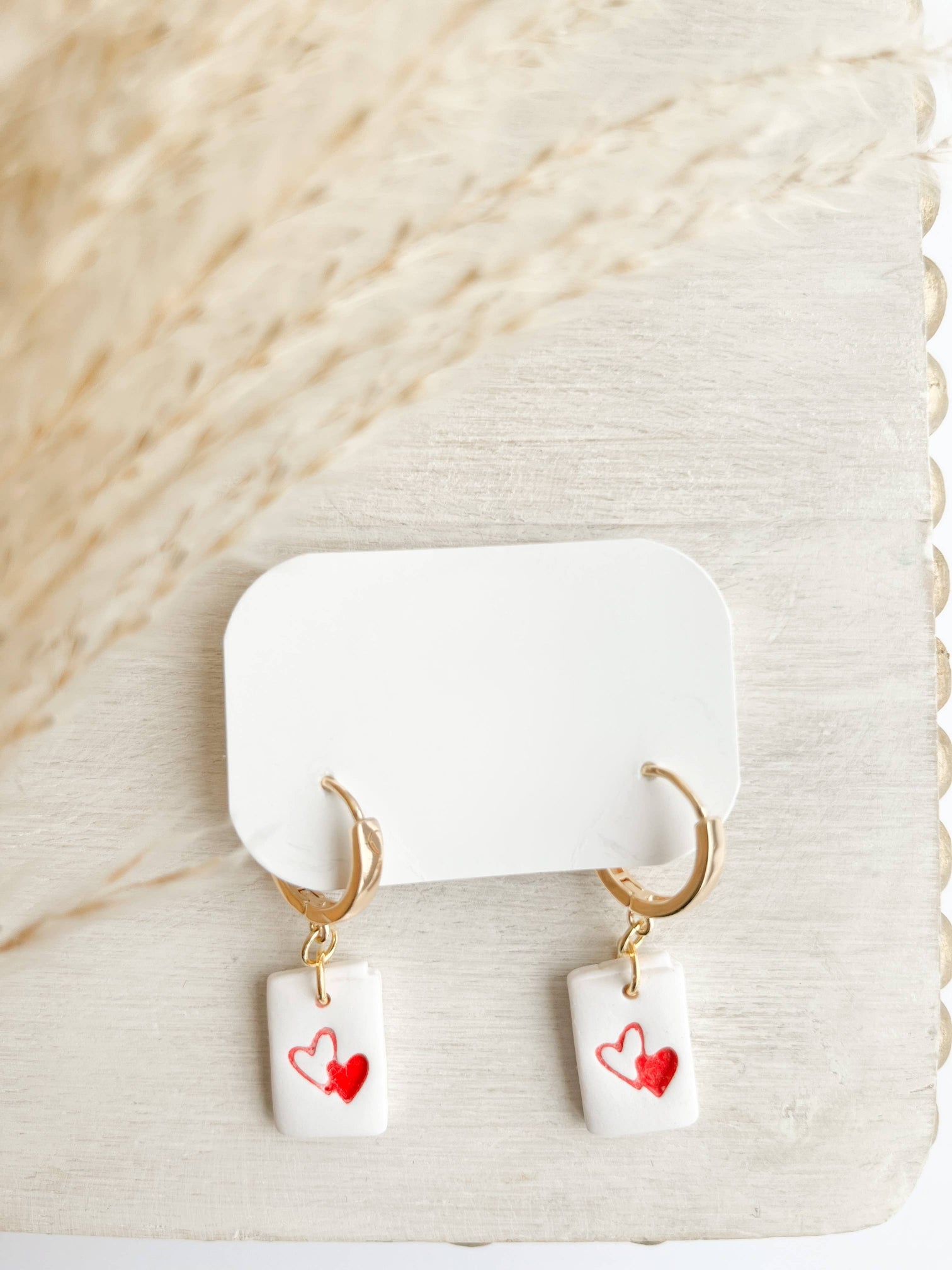 Pair of Love Letter Huggie Earrings, featuring handmade white clay rectangles with red heart designs, hanging from gold huggie hoops